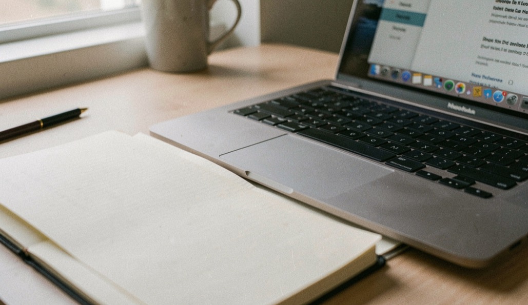 MacBook keyboard edge near paper ledger beneath window light on tidy desk